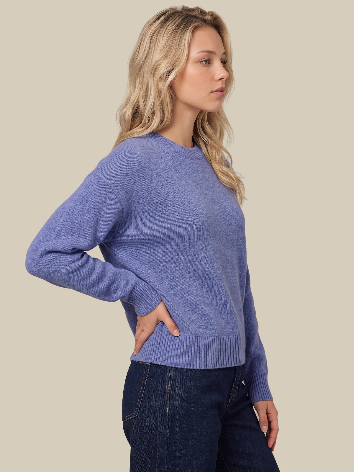 A woman wearing a heathered periwinkle blue crewneck knit sweater and dark blue jeans, standing against a solid beige background.