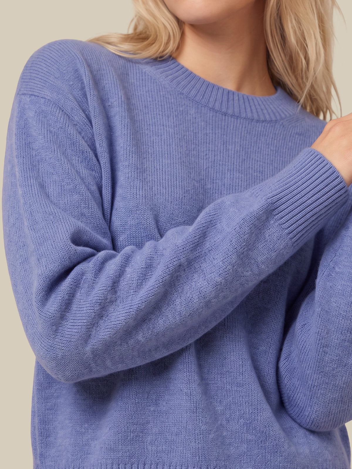 A close-up of a person wearing a ribbed knit crewneck sweater in a dusty periwinkle blue color, with blonde hair visible against a plain beige background.