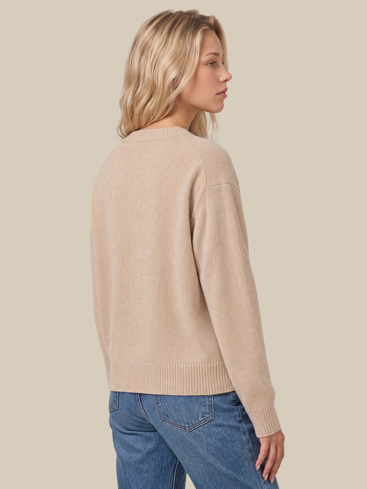 A woman shown from the back wearing a light beige knit sweater and blue jeans against a solid light beige background.