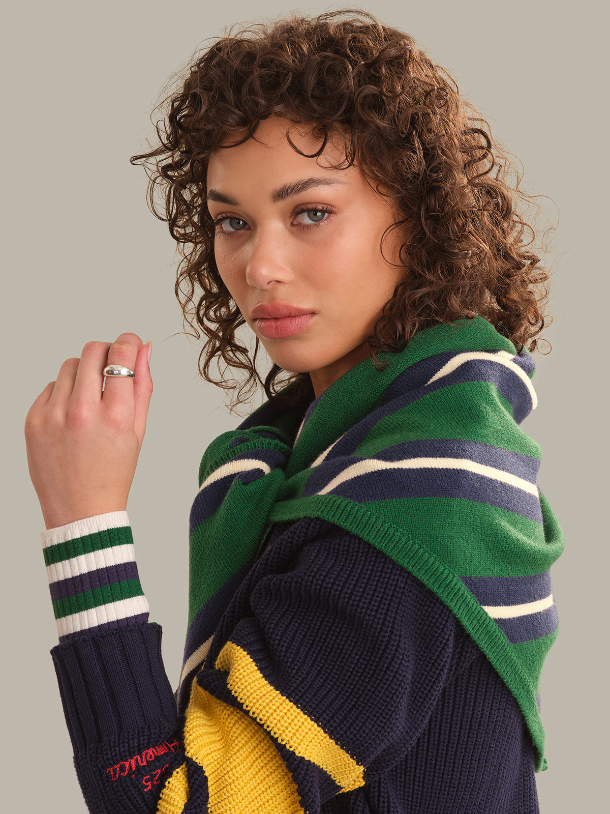A woman with curly brown hair wearing a navy blue knit sweater with yellow and green striped accents, a green striped knit shawl, and a green striped knit cuff, posed against a solid gray background.