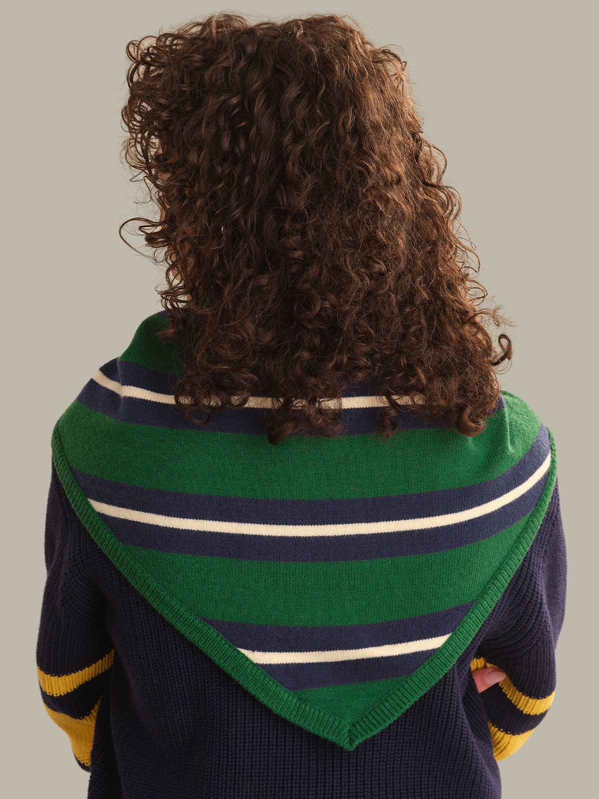 Back view of a person with curly brown hair wearing a green knit triangular shawl with navy and cream horizontal stripes over a navy sweater with yellow sleeve stripes, against a solid gray background.