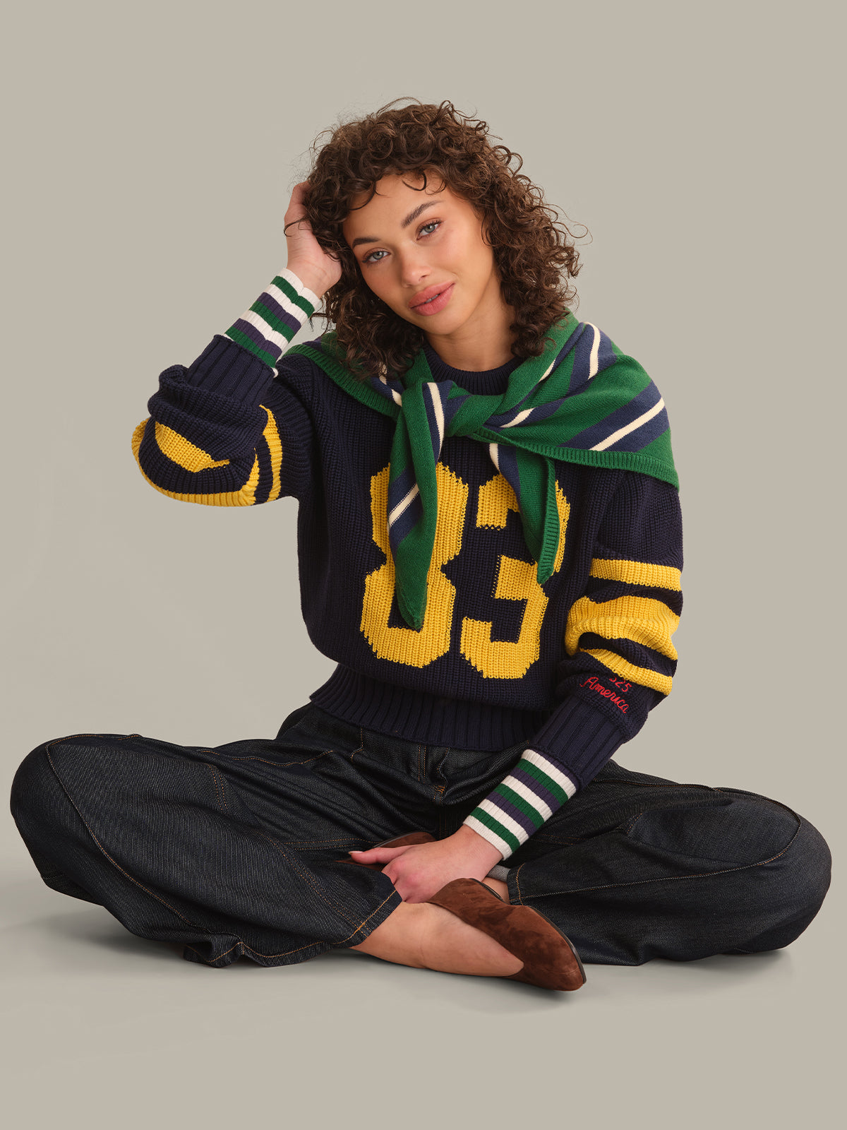 A model sits cross-legged wearing a navy blue knit sweater with yellow and green striped details, a green knit scarf, and dark pants against a solid gray background.