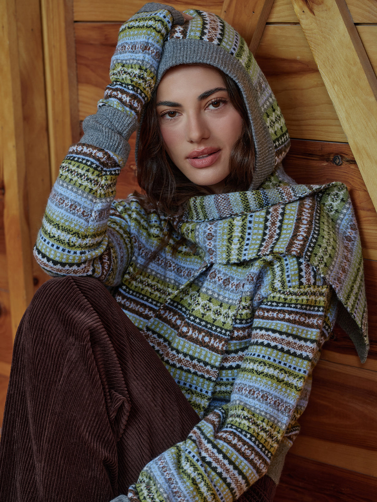 A woman wearing a knitted multicolor patterned hoodie with a large draped collar, matching patterned gloves, and brown corduroy pants, posed against a wooden wall.