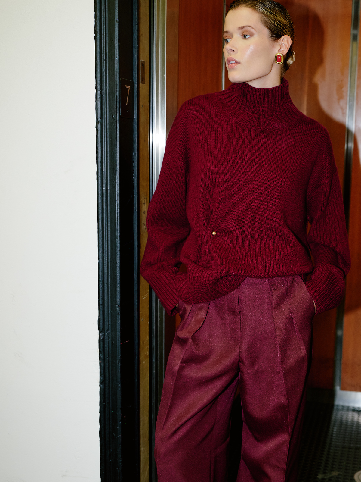 A woman wearing a deep burgundy ribbed turtleneck sweater and matching satin trousers, standing in a doorway.