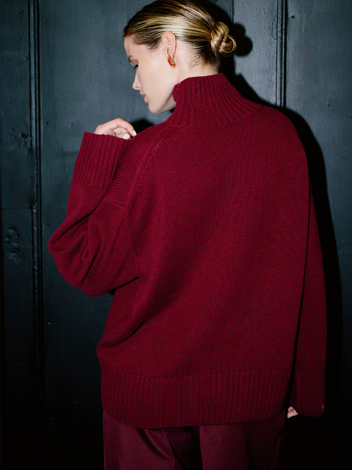 A woman wearing an oversized burgundy ribbed knit turtleneck sweater, shown from the back against a dark textured wall.