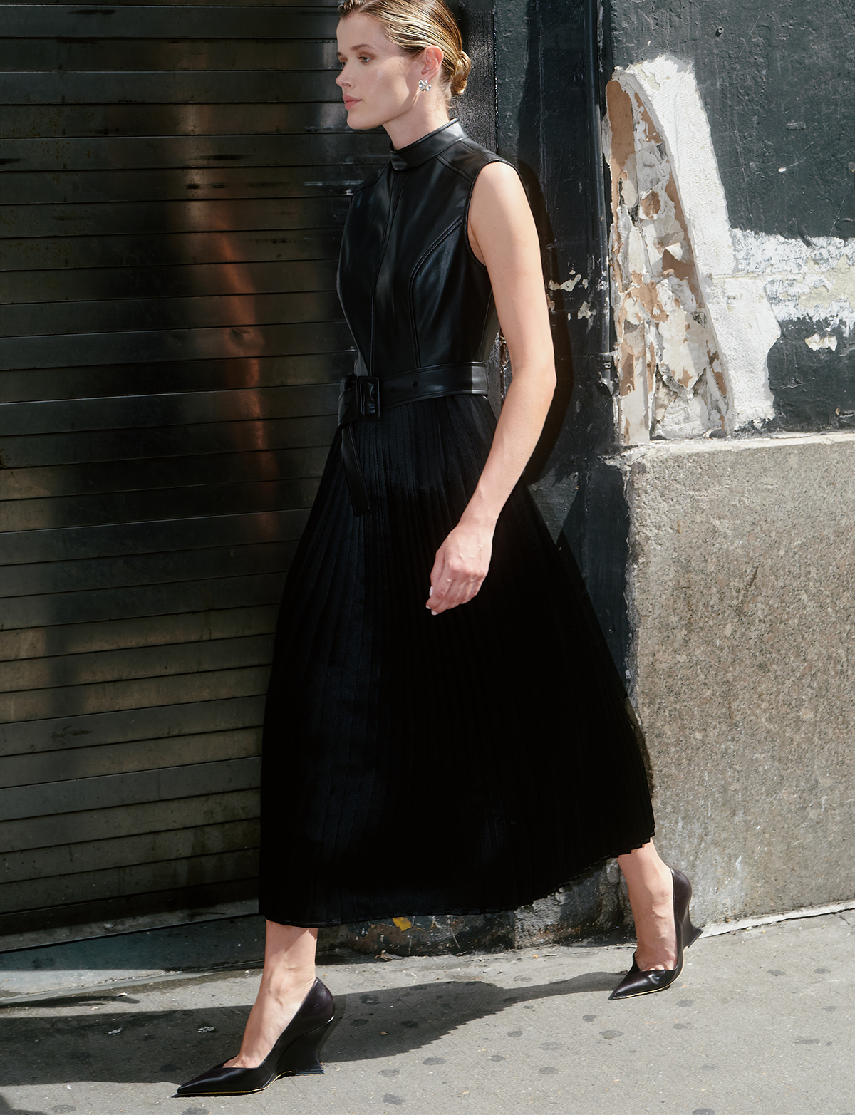 A woman models a sleeveless black pleated midi dress with a high mock neck and a matching black belt, walking outdoors against a dark textured wall.