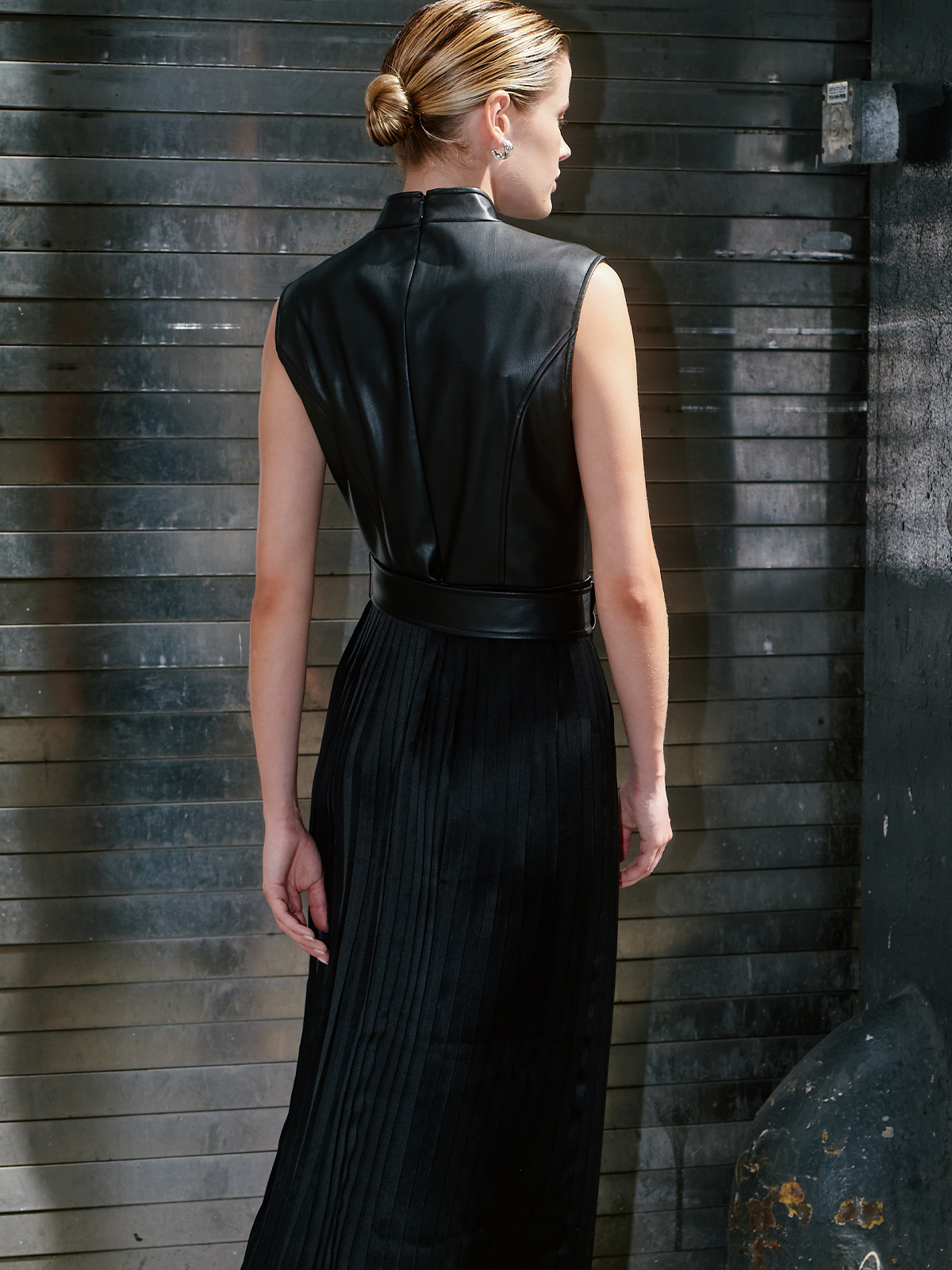 A woman shown from the back wearing a sleeveless black faux-leather vest and matching pleated black maxi skirt, standing against a dark textured wooden wall.