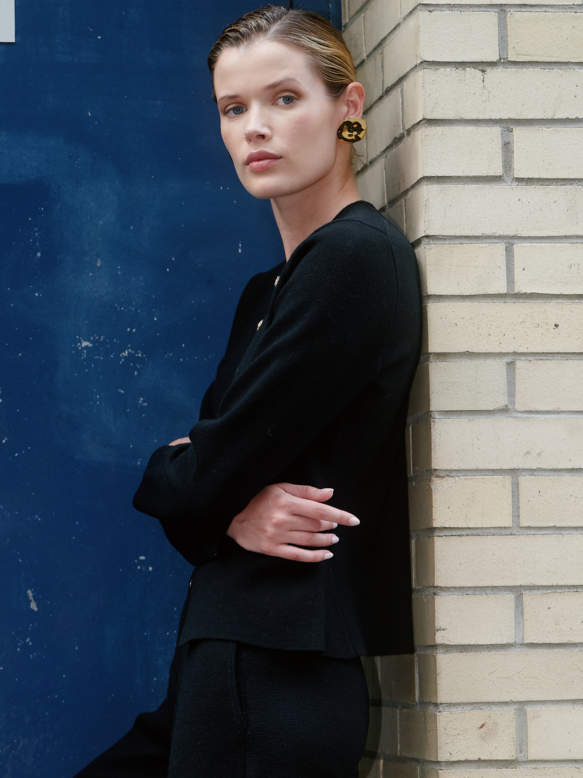 A woman with blonde hair pulled back wears a black tailored blazer and matching bottoms, leaning against a beige brick wall next to a dark blue wall. She is wearing a gold and black patterned earring.