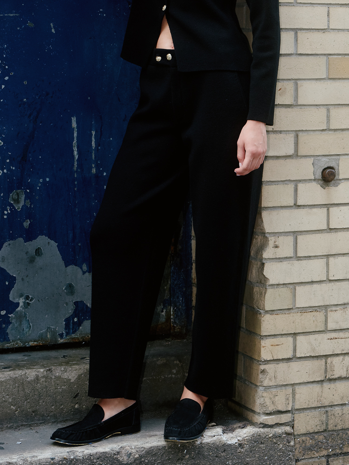 A person wearing black tailored trousers and a black cropped jacket, standing on concrete beside a blue painted wall and a light brick wall.