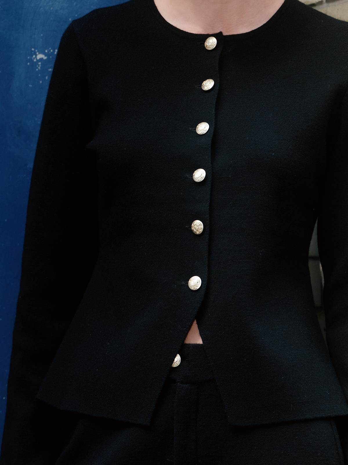 Close-up of a black tailored jacket with a round neckline and a vertical row of decorative metallic buttons down the front, worn by a model against a blue wall.