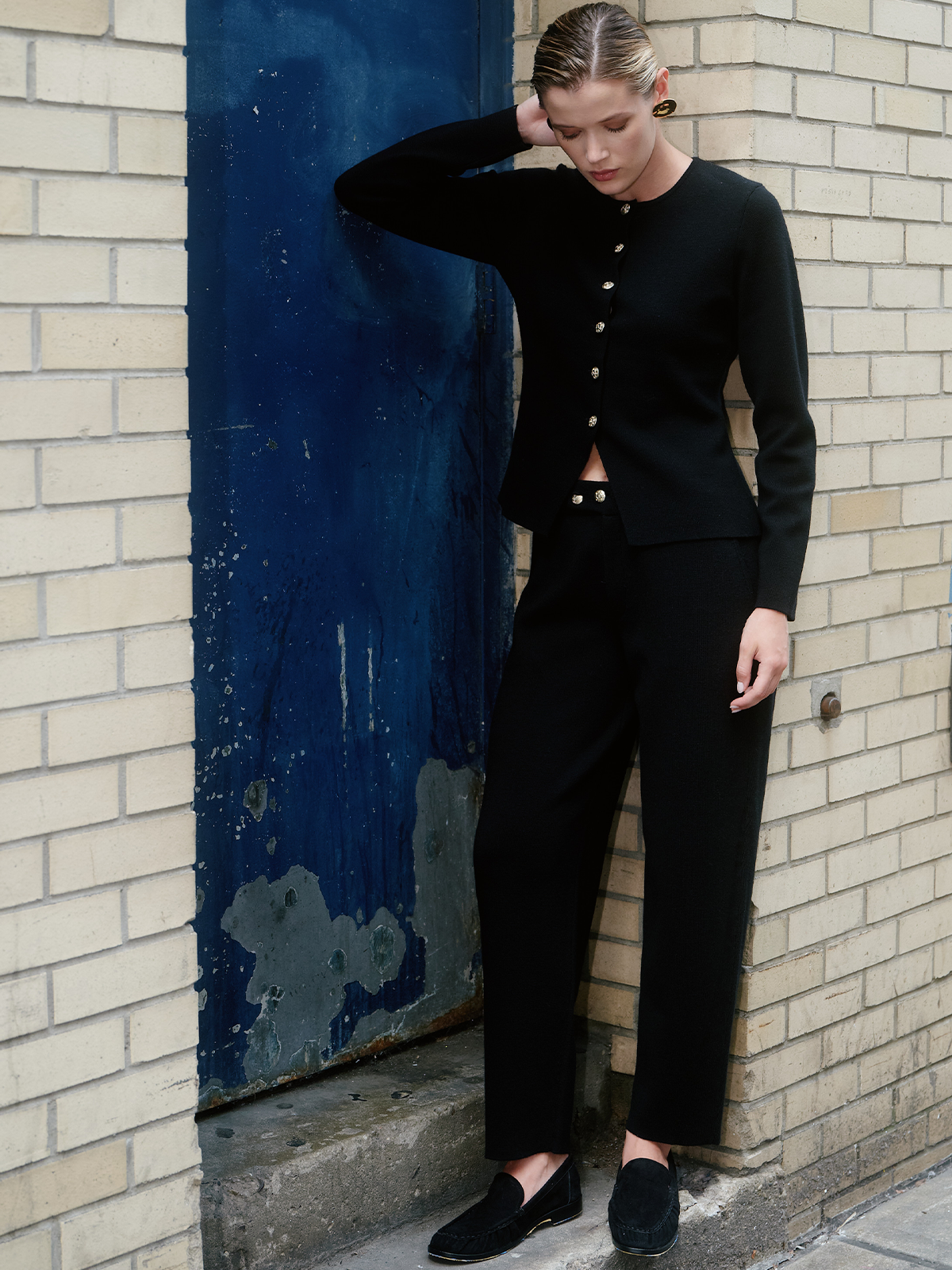 A woman models a black two-piece knit set consisting of a long-sleeve button-front top and matching cropped trousers, paired with black loafers, standing against a brick wall and a blue door.