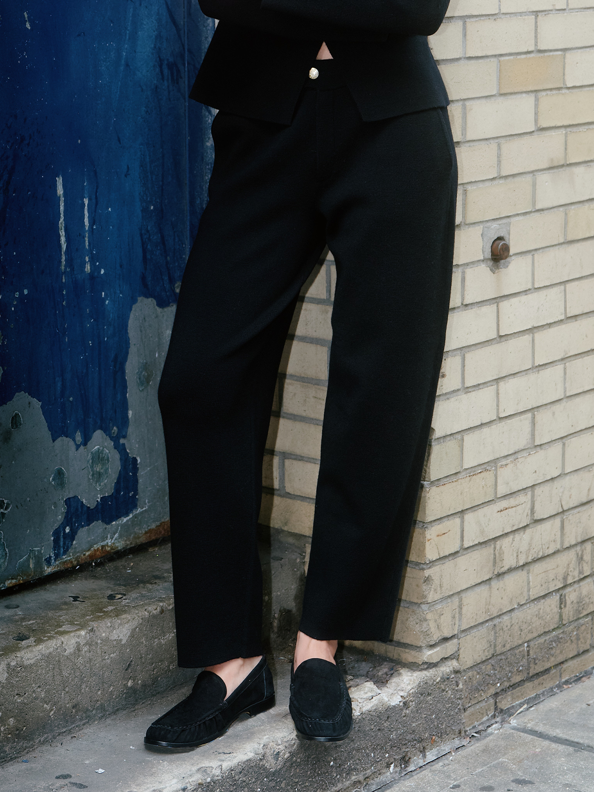 A person wearing black tailored trousers, a black blazer, and black slip-on loafers, standing on a city sidewalk against a blue metal door and beige brick wall.