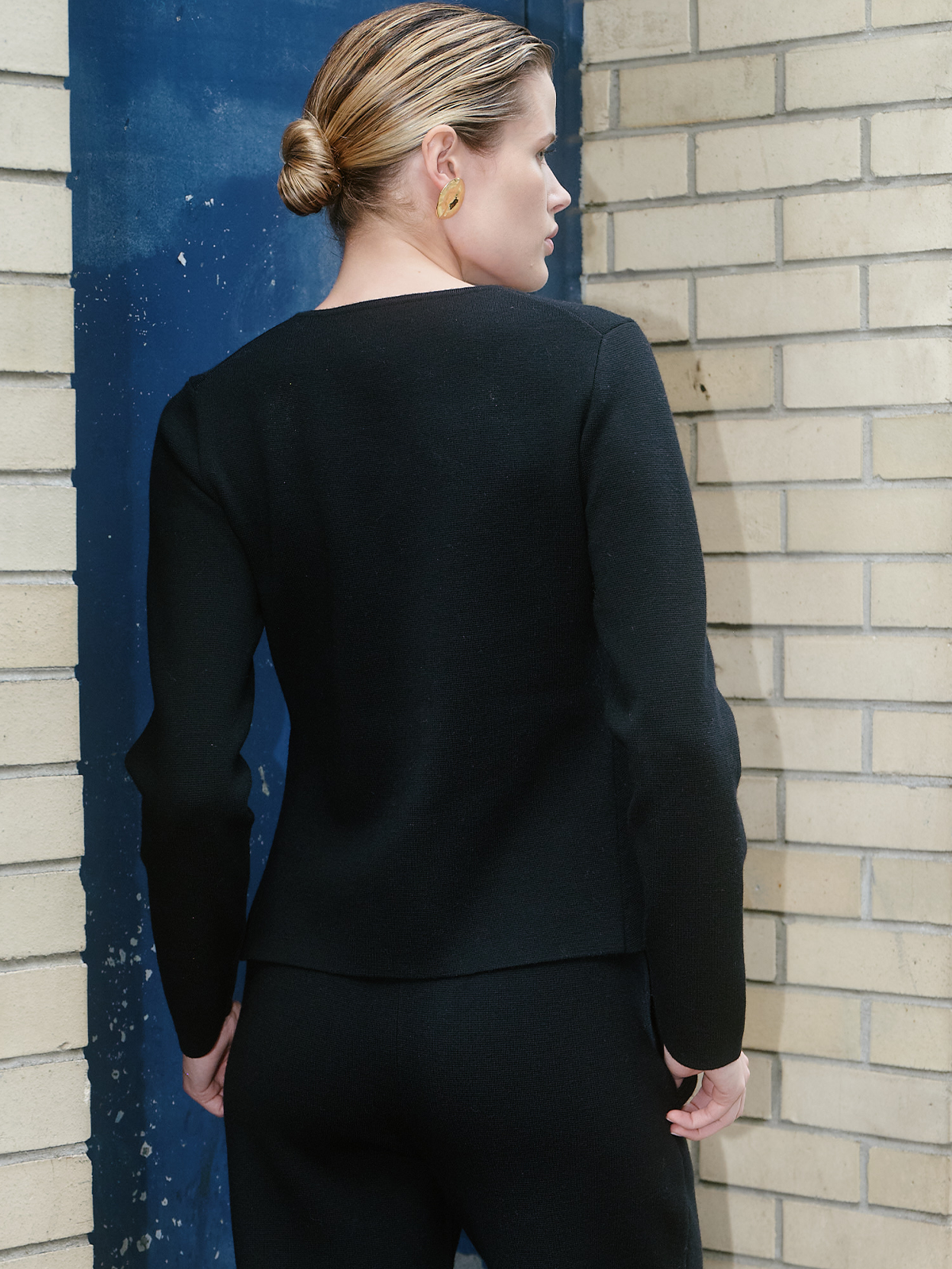 A woman shown from the back wearing a black long-sleeve knit top and matching black pants, standing against a beige brick wall and a blue door frame.
