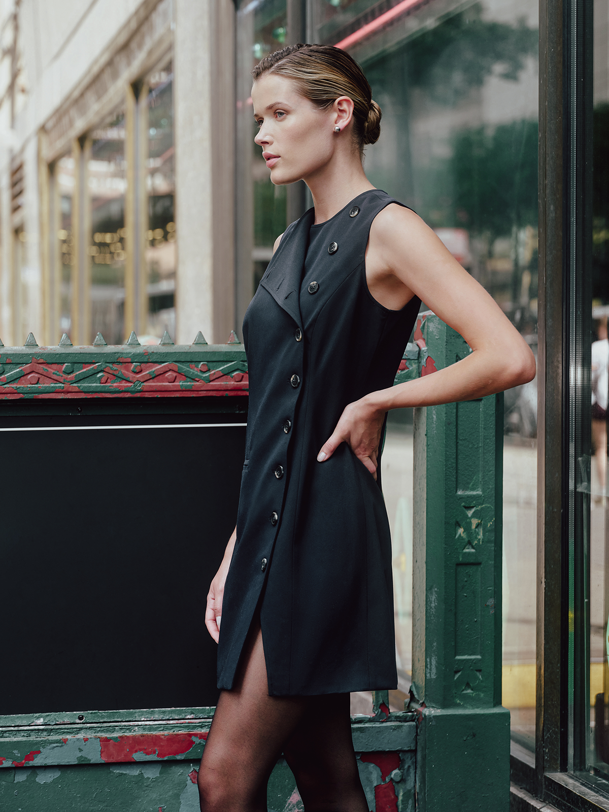 A woman models a sleeveless navy mini dress with a buttoned collar and front slit, standing next to a green decorative metal structure in an urban setting.
