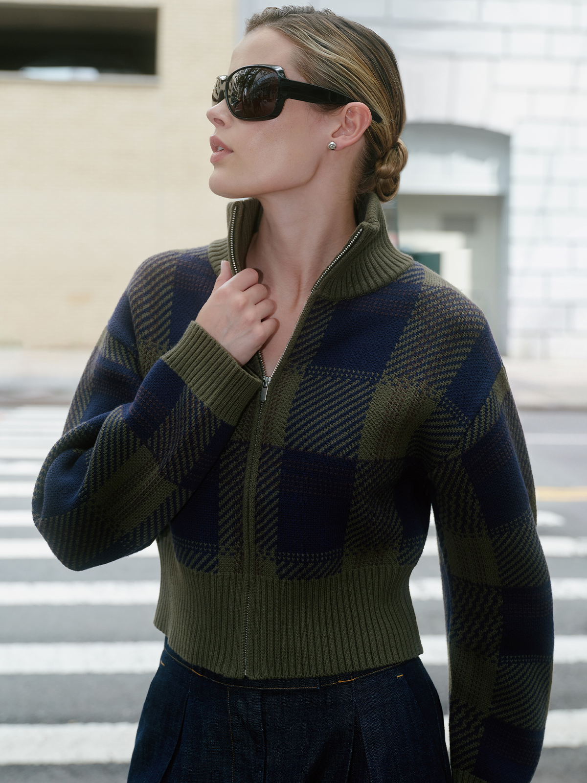 A woman wearing a cropped zip-front knit sweater with a green and navy plaid pattern, styled with dark sunglasses and a dark skirt, standing outdoors on a street crosswalk.