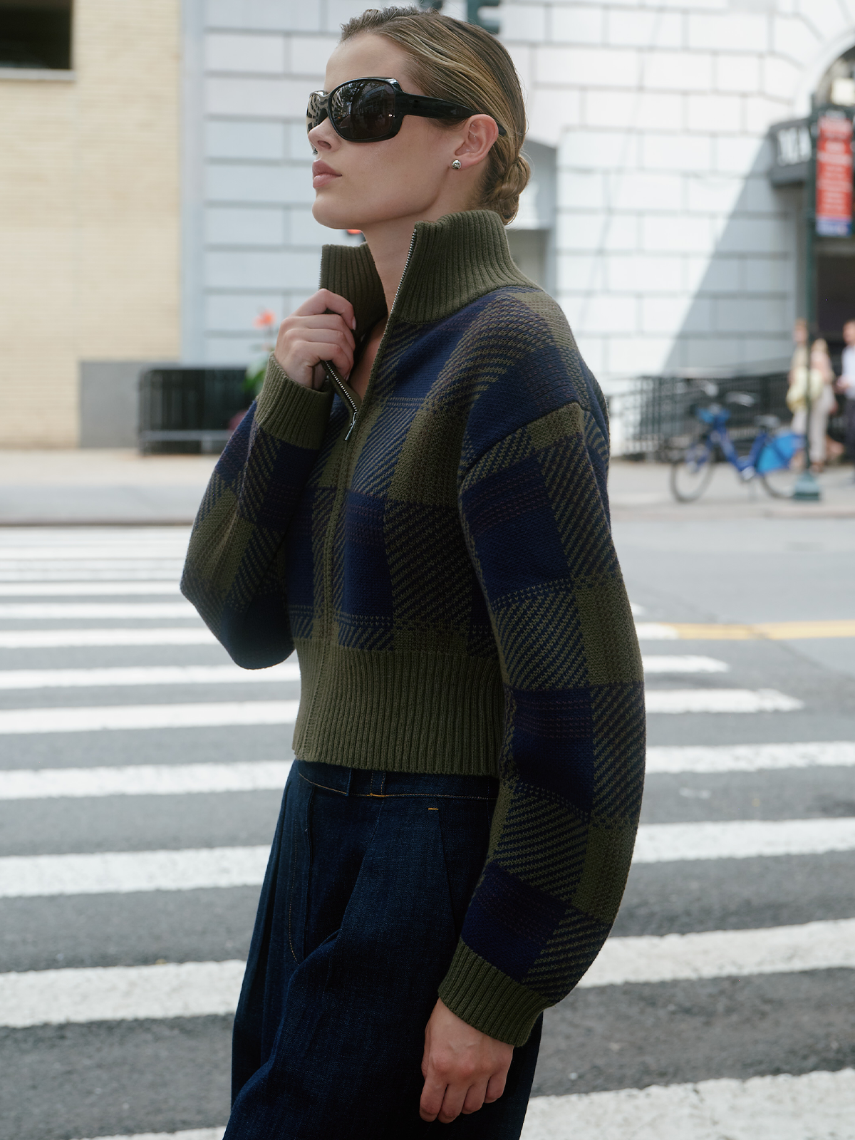 A woman wearing a green and navy blue checkered quarter-zip knit sweater and dark pants, standing on a city crosswalk.