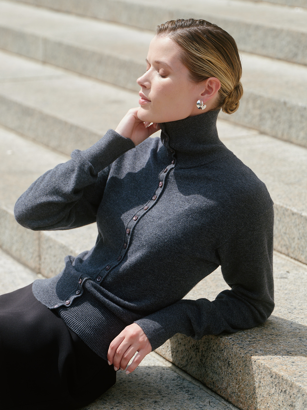 A woman wearing a dark gray turtleneck cardigan with decorative buttons, sitting on stone steps outdoors.