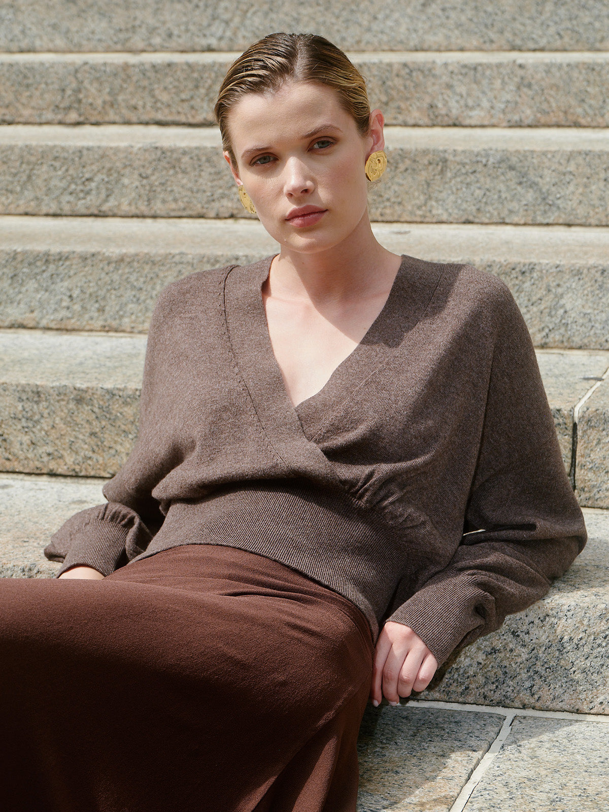 A woman sits on stone steps wearing a brown knit wrap-style sweater and matching brown skirt, with gold earrings.