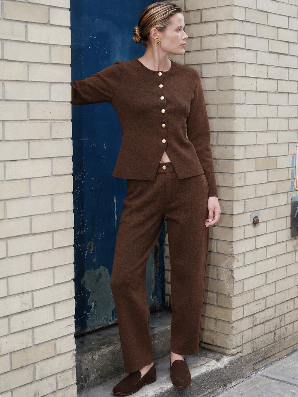 A woman models a matching brown knit two-piece set consisting of a cropped long-sleeve cardigan with decorative buttons and matching wide-leg trousers, standing against a beige brick wall and a blue door.