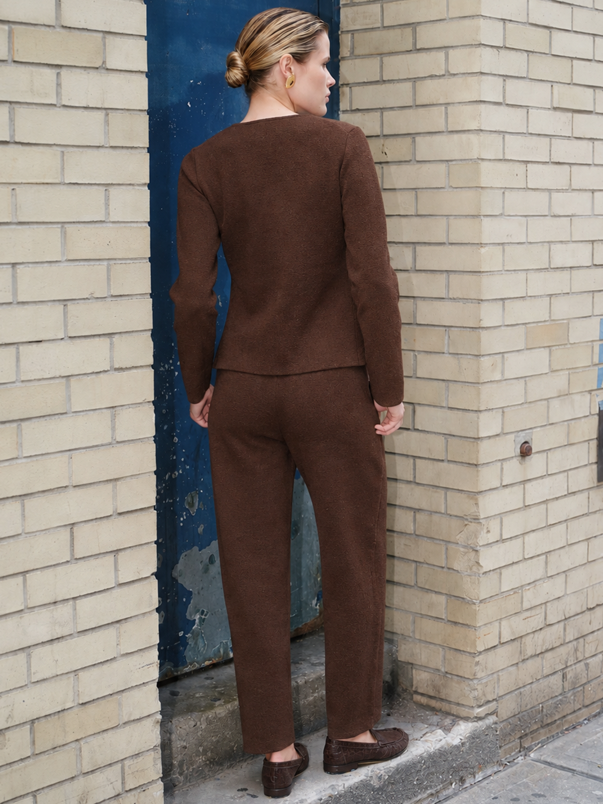 A woman stands with her back to the camera wearing a matching brown knit long-sleeve top and trousers set, positioned in front of a beige brick wall and a blue door.